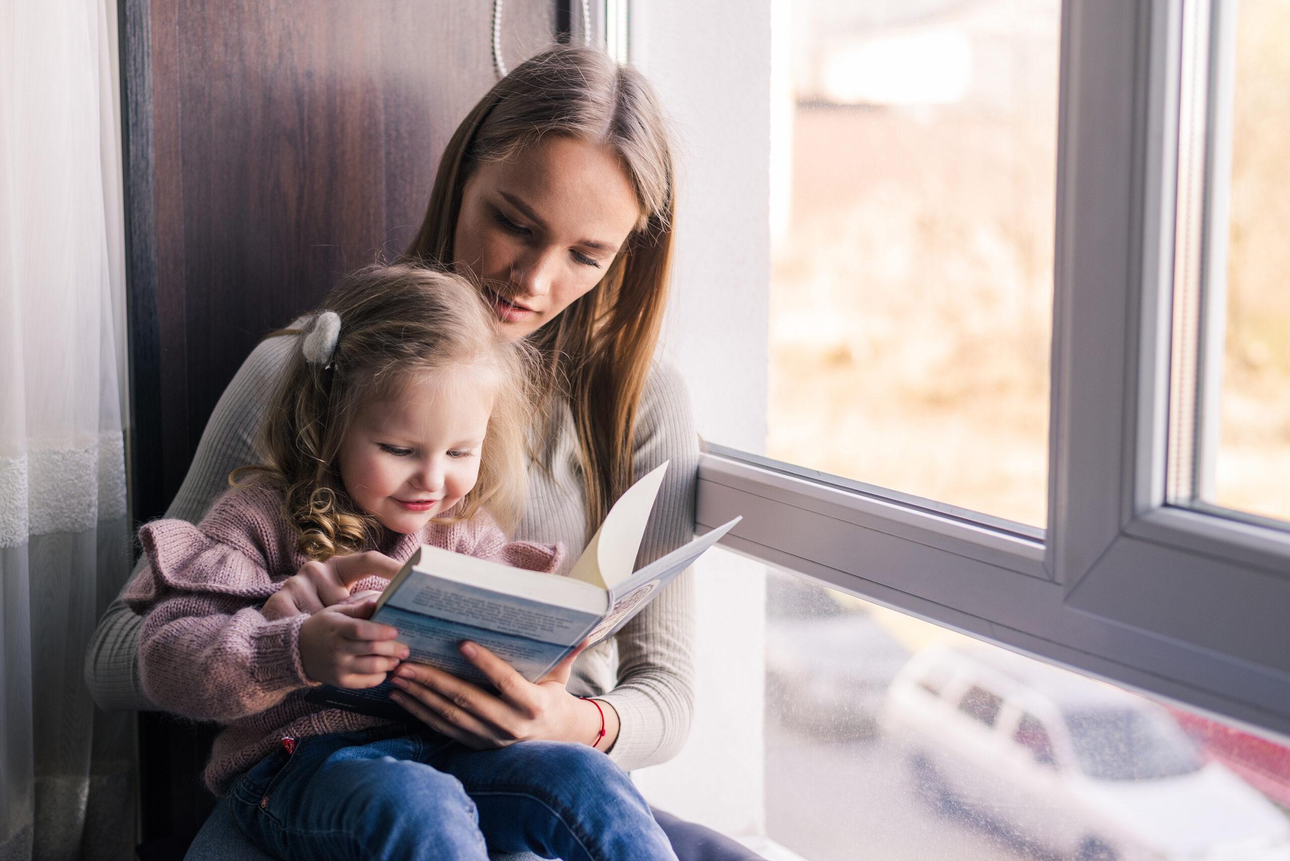 Happy mother and little daughter reading book, sitting on comfortable couch in living room, smiling mum teaching little girl child, family spending weekend at home together, children education